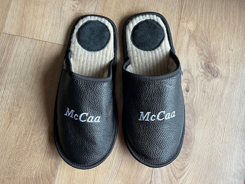 May include: A pair of black leather slippers with white embroidered text that reads "McCaа". The slippers have a beige fleece lining.