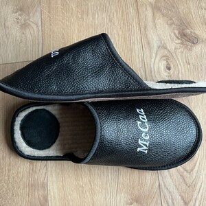May include: A pair of black leather slippers with a white embroidered name "McCa" on one slipper. The slippers have a soft, fuzzy lining.