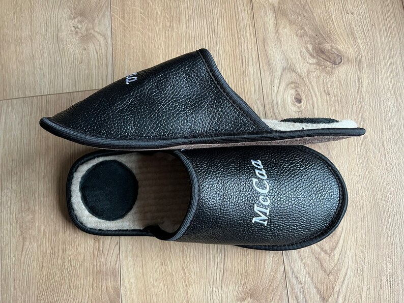 May include: A pair of black leather slippers with a white embroidered name "McCa" on one slipper. The slippers have a soft, fuzzy lining.