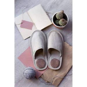 Op de afbeelding: Een paar grijze linnen slippers met bruine su&egrave;de inlegzolen. De slippers liggen op een bruine papieroppervlak met een bol wol en een roze papiertje.