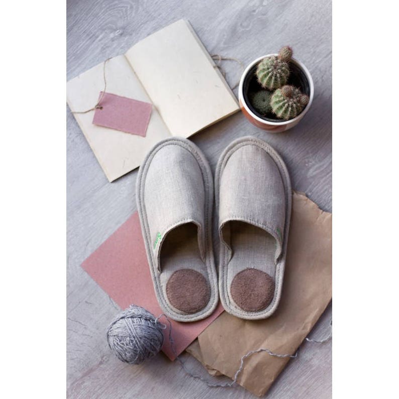 Op de afbeelding: Een paar grijze linnen slippers met bruine su&egrave;de inlegzolen. De slippers liggen op een bruine papieroppervlak met een bol wol en een roze papiertje.