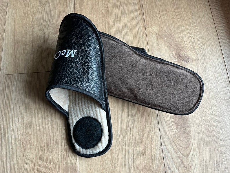 May include: A pair of black leather slippers with a brown fleece lining. The slippers have a white embroidered logo that says "MCC".