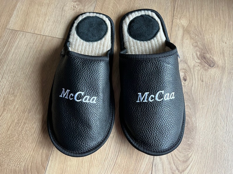 May include: A pair of black leather slippers with white embroidered text that reads "McCaа". The slippers have a soft, beige lining.