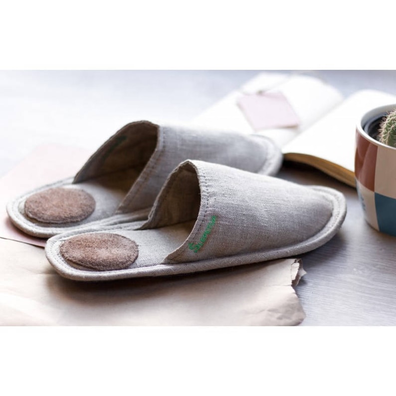 Op de afbeelding: Een paar lichtgrijze linnen slippers met bruine su&egrave;de zolen. De slippers hebben een eenvoudig ontwerp en zijn perfect om in huis te dragen.