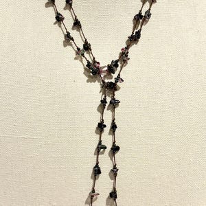 May include: A long lariat-style necklace featuring two strands of dark brown cord knotted at intervals. The cord is adorned with small, irregular, tumbled gemstone beads in shades of deep purple, black, and translucent pink, ending in two dangling sections.