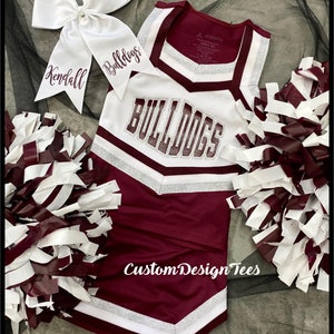 May include: A maroon and white cheerleading uniform featuring the word BULLDOGS in glittery lettering, a matching white hair bow with the text Kendall Bulldogs, and two maroon and white pom-poms. The uniform includes silver glitter accents.
