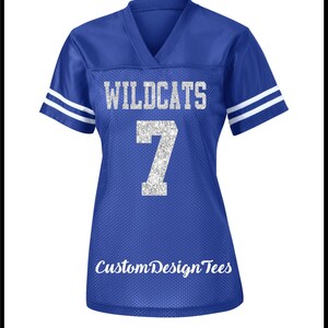 May include: A royal blue V-neck jersey with short sleeves featuring two white stripes on each sleeve. The front displays the silver glitter text WILDCATS above the number 7 in silver glitter. The text CustomDesignTees appears at the bottom.