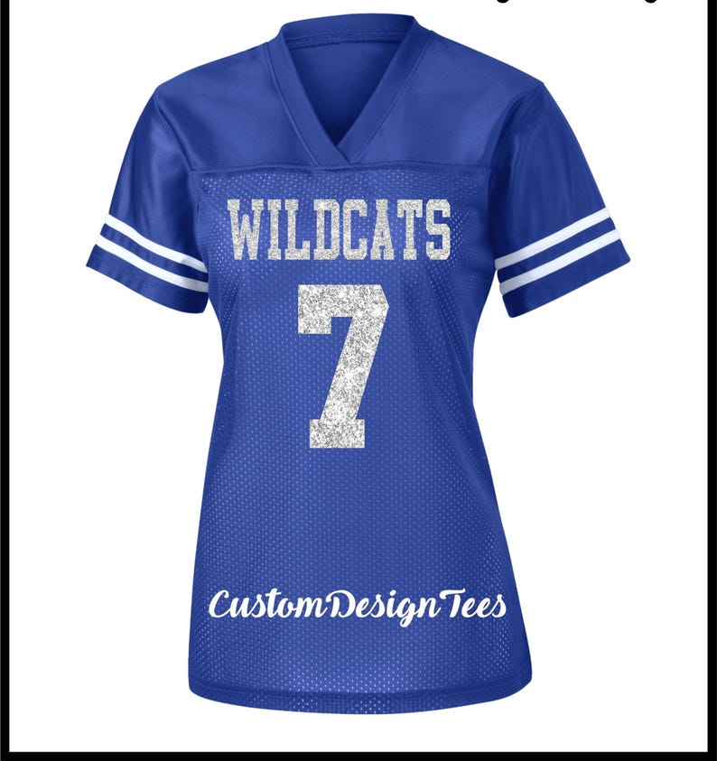 May include: A royal blue V-neck jersey with short sleeves featuring two white stripes on each sleeve. The front displays the silver glitter text WILDCATS above the number 7 in silver glitter. The text CustomDesignTees appears at the bottom.