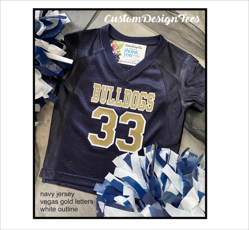 May include: Navy blue jersey with "BULLDOGS" and the number "33" in Vegas gold letters with a white outline. The jersey has a V-neck design. The image also includes pom-poms with navy and white colors.