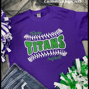 May include: A purple t-shirt with a green glitter design that reads "Texas Titans Softball." The design is in the shape of a baseball. The shirt is paired with denim jeans and pom-poms.