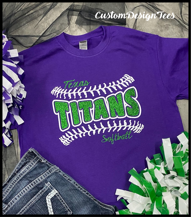 May include: A purple t-shirt with a green glitter design that reads "Texas Titans Softball." The design is in the shape of a baseball. The shirt is paired with denim jeans and pom-poms.