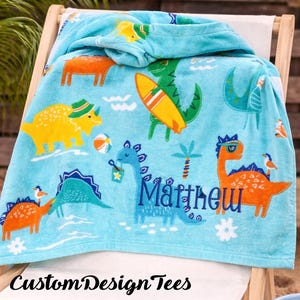 Pode incluir: Uma toalha de praia azul clara com um tema de dinossauros. A toalha apresenta v&aacute;rias ilustra&ccedil;&otilde;es coloridas de dinossauros, uma prancha de surf e o nome "Matthew" em escrita azul. O texto "Custom Design Tees" est&aacute; na parte inferior.