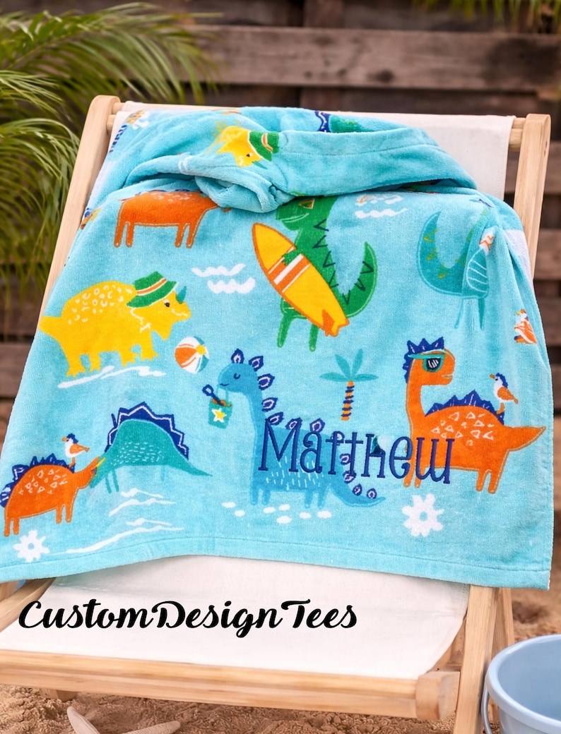 Pode incluir: Uma toalha de praia azul clara com um tema de dinossauros. A toalha apresenta v&aacute;rias ilustra&ccedil;&otilde;es coloridas de dinossauros, uma prancha de surf e o nome "Matthew" em escrita azul. O texto "Custom Design Tees" est&aacute; na parte inferior.