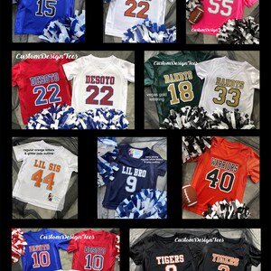 May include: Collection of youth sports jerseys in various colors, including blue, white, red, green, orange, and black. Each jersey features team names, numbers, and pom-poms. The jerseys are for teams like Bobcats, Bulldogs, Wildcats, and Tigers.