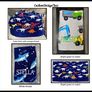 May include: Four personalized towels and a drawstring bag are displayed. The bag is navy blue with dinosaur designs and the name "BRODY" in orange. One towel has construction vehicles and the name "PRESTON" in green. Another has sharks and the name "STELLA". The last towel has dinosaurs and the name "SALEM".