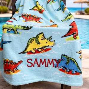 May include: A light blue beach towel featuring colorful cartoon dinosaurs. The towel has a repeating pattern of various dinosaurs in yellow, red, green, and blue. The name "SAMMY" is embroidered in red. The text "Custom Design Tees" is at the bottom.
