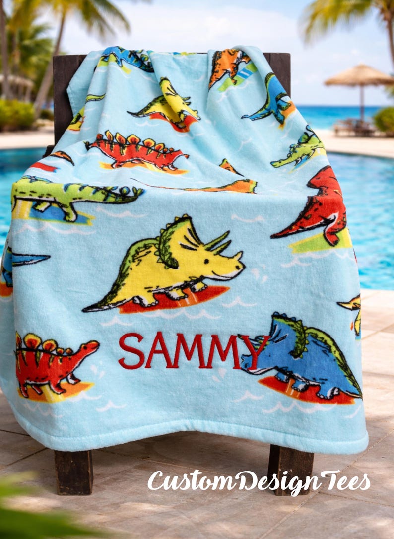 May include: A light blue beach towel featuring colorful cartoon dinosaurs. The towel has a repeating pattern of various dinosaurs in yellow, red, green, and blue. The name "SAMMY" is embroidered in red. The text "Custom Design Tees" is at the bottom.