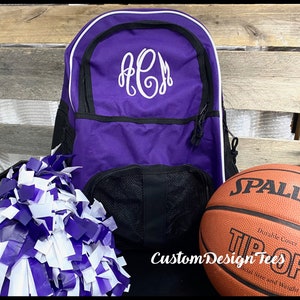 May include: Purple and black backpack with white trim and a white monogram "ACJ" on the front. A white and purple pom pom and a brown basketball with the word "SPALDING" are in the foreground.