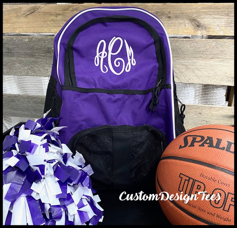 May include: Purple and black backpack with white trim and a white monogram "ACJ" on the front. A white and purple pom pom and a brown basketball with the word "SPALDING" are in the foreground.