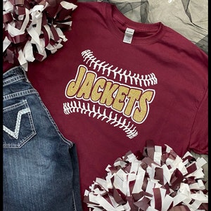May include: A maroon t-shirt with the word "JACKETS" in gold glitter, outlined with white baseball stitching. The shirt is paired with blue jeans and maroon and white pom-poms. The text "Custom Design Tees" is in the top right corner.