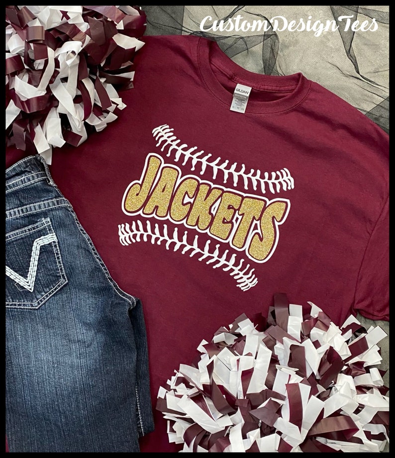 May include: A maroon t-shirt with the word "JACKETS" in gold glitter, outlined with white baseball stitching. The shirt is paired with blue jeans and maroon and white pom-poms. The text "Custom Design Tees" is in the top right corner.