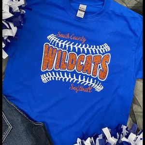 May include: Royal blue t-shirt with a softball design. The design features the words "South County Wildcats Softball" in white and orange glitter. The shirt is paired with denim jeans and blue and white pom-poms. The text "Custom Design Tees" is at the top.