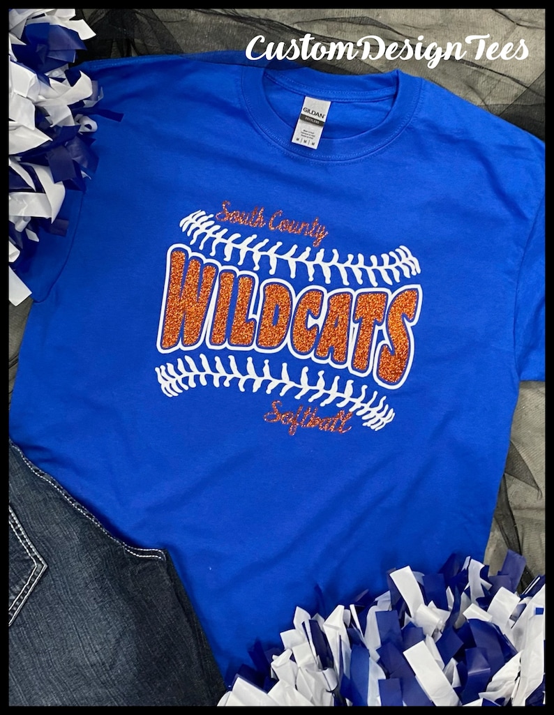 May include: Royal blue t-shirt with a softball design. The design features the words "South County Wildcats Softball" in white and orange glitter. The shirt is paired with denim jeans and blue and white pom-poms. The text "Custom Design Tees" is at the top.