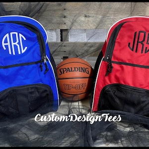 May include: Two personalized sports bags, one blue and one red, with white trim and black mesh pockets. The bags are monogrammed with the initials "ARC" and "JRS". A basketball with the Spalding logo and "Tip-Off" text is in the center.