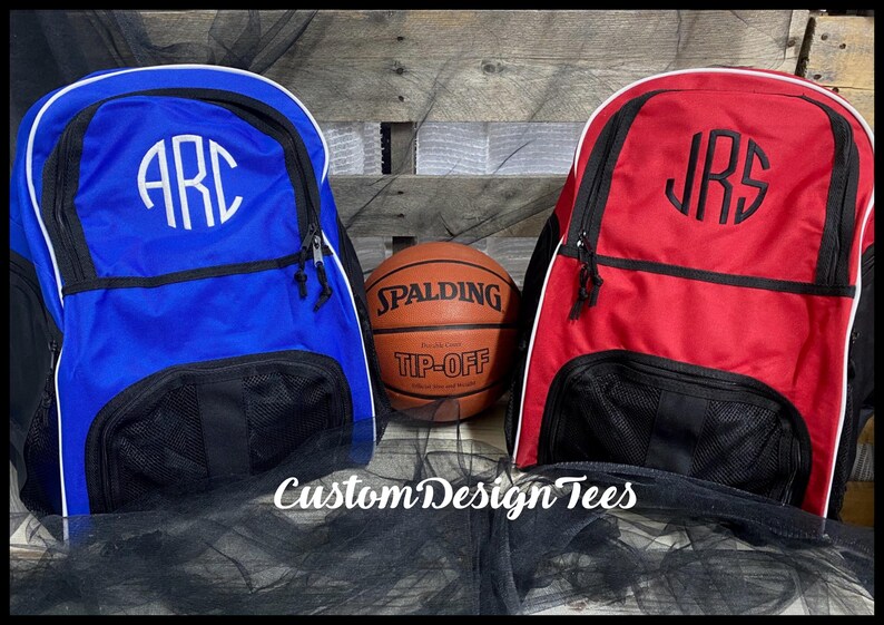 May include: Two personalized sports bags, one blue and one red, with white trim and black mesh pockets. The bags are monogrammed with the initials "ARC" and "JRS". A basketball with the Spalding logo and "Tip-Off" text is in the center.