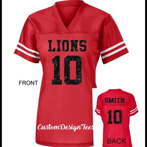 May include: A red V-neck jersey with short sleeves featuring two white stripes on each sleeve. The front displays the word LIONS and the number 10 in black glitter. The back shows the name SMITH and the number 10 in black glitter text.