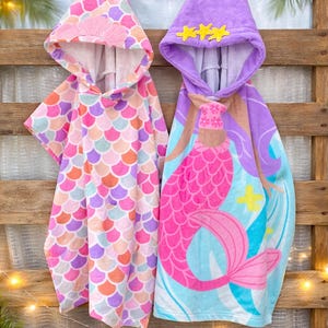 Pode incluir: Duas toalhas de praia com capuz penduradas em um fundo de madeira. Uma toalha apresenta um padr&atilde;o de escamas em rosa, laranja e roxo. A outra toalha tem um design de sereia em rosa, roxo e azul. O texto "Custom Design Tees" &eacute; vis&iacute;vel.
