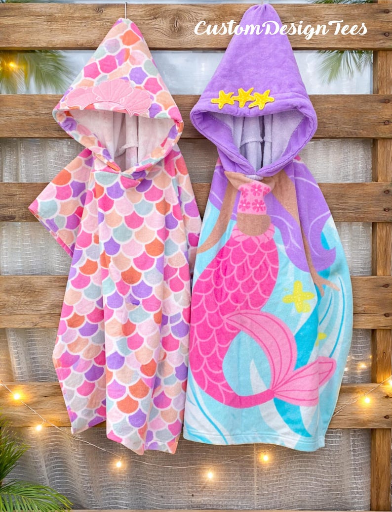 Pode incluir: Duas toalhas de praia com capuz penduradas em um fundo de madeira. Uma toalha apresenta um padr&atilde;o de escamas em rosa, laranja e roxo. A outra toalha tem um design de sereia em rosa, roxo e azul. O texto "Custom Design Tees" &eacute; vis&iacute;vel.