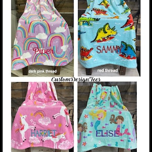 May include: Four personalized children's towels with embroidered names. The towels feature colorful designs: rainbows, dinosaurs, unicorns, and mermaids. The names Piper, Sammy, Harriet, and Elise are embroidered in dark pink, red, aqua, and dark purple thread.