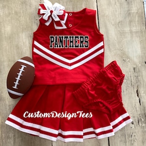 May include: A red cheerleader outfit with white accents, including a top with "PANTHERS" in black, a pleated skirt, and matching shorts. A football and a bow complete the ensemble. The text "CustomDesignTees" is visible.