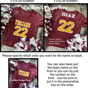 May include: Maroon jersey with yellow gold lettering and a white outline. The front displays "TROJANS" and the number "22". The back shows "DIAZ" and "22". The image also shows a jersey with "TITANS" on the front. Pom-poms are in the foreground.
