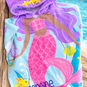 Pode incluir: Uma toalha de praia com capuz com um design de sereia. A toalha &eacute; azul clara com uma sereia rosa, cabelo roxo e detalhes amarelos. O capuz &eacute; roxo com enfeites de estrela amarelos. O nome "Jasmine" est&aacute; impresso.