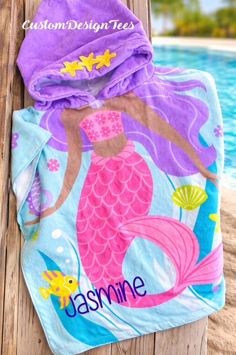 Pode incluir: Uma toalha de praia com capuz com um design de sereia. A toalha &eacute; azul clara com uma sereia rosa, cabelo roxo e detalhes amarelos. O capuz &eacute; roxo com enfeites de estrela amarelos. O nome "Jasmine" est&aacute; impresso.
