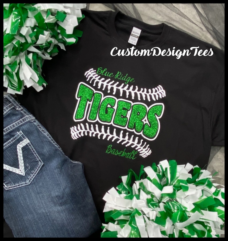May include: Black t-shirt with a baseball design featuring the text "Blue Ridge Tigers Baseball" in green glitter. The shirt is paired with blue jeans and green and white pom-poms. The text "Custom Design Tees" is also visible.
