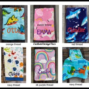May include: Collection of personalized beach towels with embroidered names. Designs include dinosaurs, sharks, sea creatures, rainbows, and unicorns. Towels are in various colors, including blue, pink, and purple. The text "Custom Design Tees" is also visible.