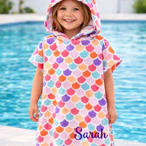 Pode incluir: Uma toalha de praia com capuz com um padr&atilde;o de escamas de sereia em tons de rosa, laranja, roxo e azul. O capuz apresenta um design de concha. O nome "Sarah" est&aacute; impresso na toalha. O texto "CustomDesignTees" tamb&eacute;m &eacute; vis&iacute;vel.