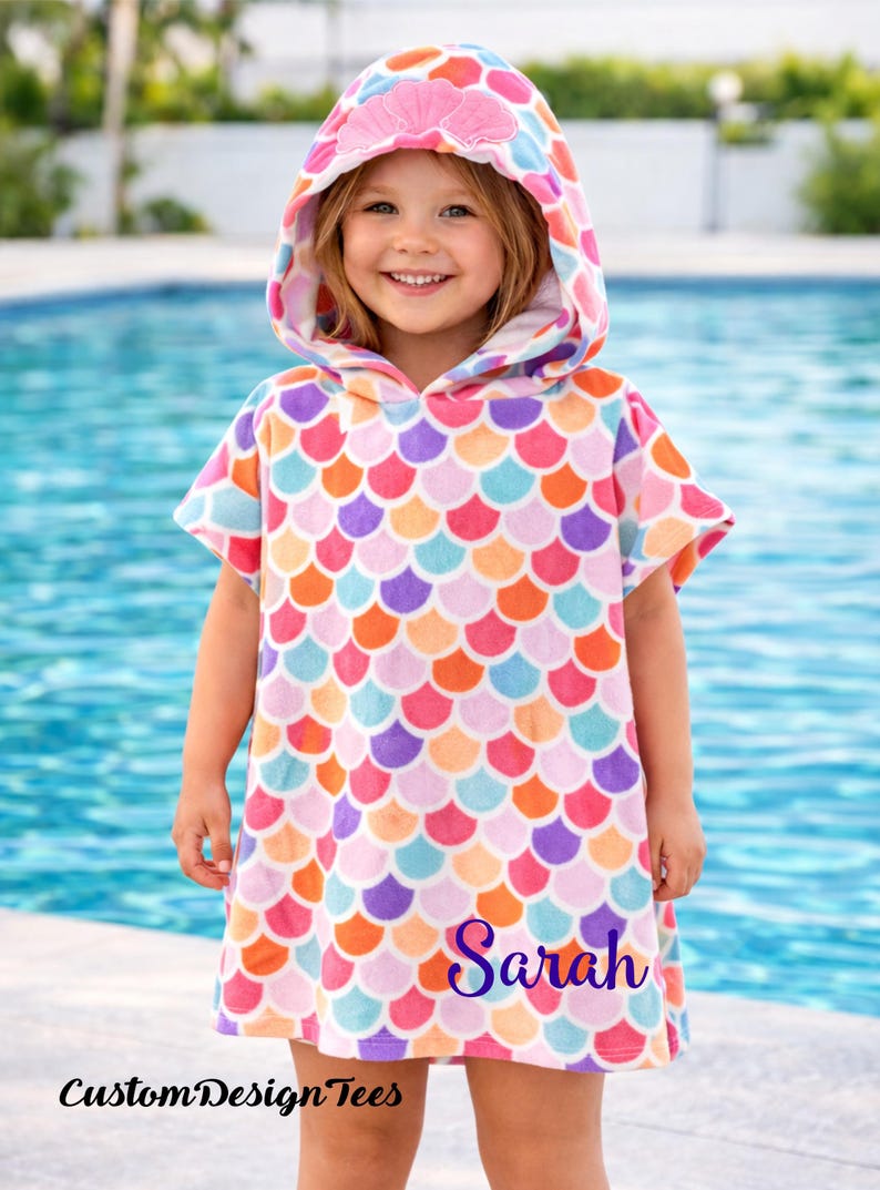 Pode incluir: Uma toalha de praia com capuz com um padr&atilde;o de escamas de sereia em tons de rosa, laranja, roxo e azul. O capuz apresenta um design de concha. O nome "Sarah" est&aacute; impresso na toalha. O texto "CustomDesignTees" tamb&eacute;m &eacute; vis&iacute;vel.