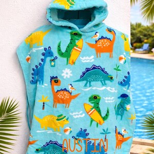 Pode incluir: Uma toalha de praia com capuz azul claro com um padr&atilde;o repetido de dinossauros de desenho animado coloridos, alguns com pranchas de surf e chap&eacute;us. O nome "Austin" &eacute; impresso em laranja e azul. O texto "Custom Design Tees" &eacute; impresso no canto superior esquerdo.