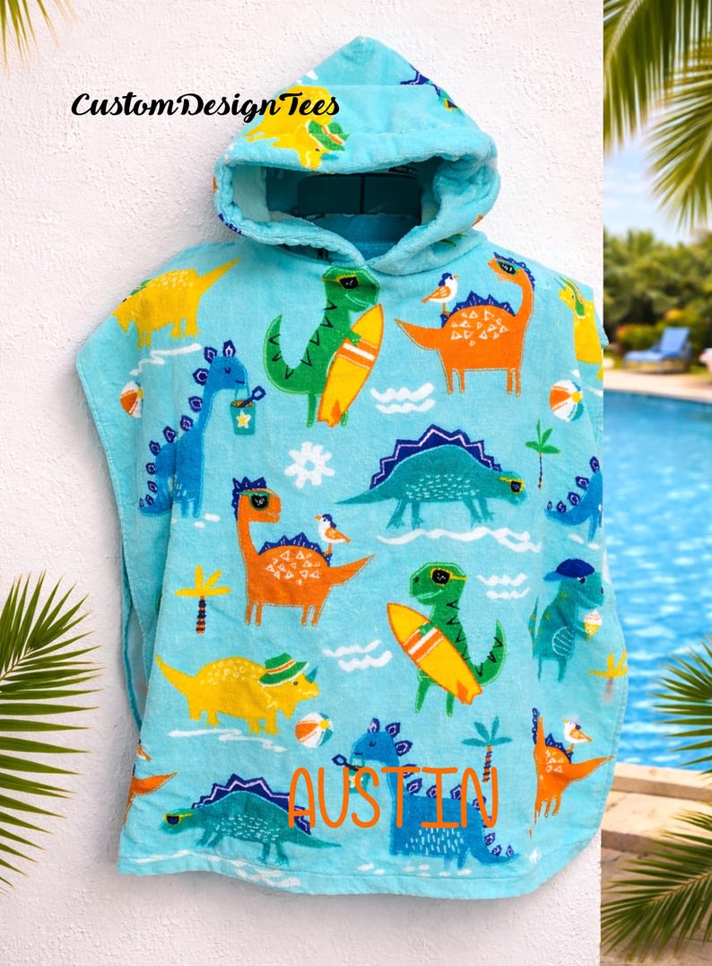 Pode incluir: Uma toalha de praia com capuz azul claro com um padr&atilde;o repetido de dinossauros de desenho animado coloridos, alguns com pranchas de surf e chap&eacute;us. O nome "Austin" &eacute; impresso em laranja e azul. O texto "Custom Design Tees" &eacute; impresso no canto superior esquerdo.
