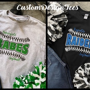May include: Two t-shirts with baseball-themed designs. One is gray with "Bakersfield BABES Softball" in green glitter. The other is black with "Reynolds RAIDERS Softball" in blue glitter. Pom-poms and jeans are also visible.