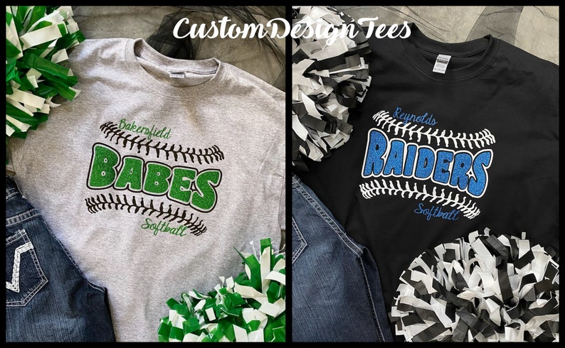 May include: Two t-shirts with baseball-themed designs. One is gray with "Bakersfield BABES Softball" in green glitter. The other is black with "Reynolds RAIDERS Softball" in blue glitter. Pom-poms and jeans are also visible.