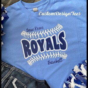 May include: A light blue t-shirt with the text "Base Knox ROYALS Baseball" in white and navy blue. The shirt features a baseball design and the brand name "CustomDesignTees" in white. Dark blue jeans and blue and white pom-poms are also in the image.