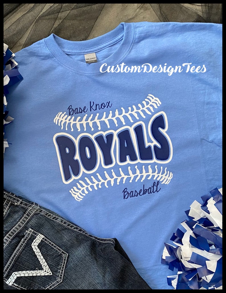 May include: A light blue t-shirt with the text "Base Knox ROYALS Baseball" in white and navy blue. The shirt features a baseball design and the brand name "CustomDesignTees" in white. Dark blue jeans and blue and white pom-poms are also in the image.