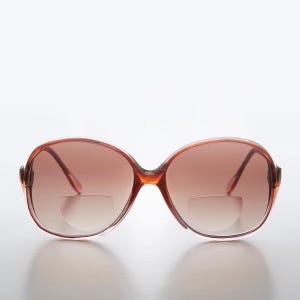 Puede incluir: Un par de gafas de lectura ovaladas de color marrón con monturas de plástico transparente. Las gafas tienen un tinte degradado marrón.