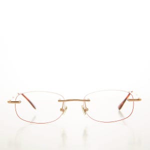 May include: Rimless spectacles with a gold-coloured bridge and temples. The lenses are oval-shaped with a thin, red-coloured outline. The spectacles have a minimalist design.