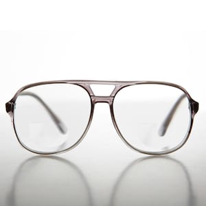 Puede incluir: Gafas de estilo aviador con montura de color lila claro. Las lentes son transparentes y la montura presenta un diseño de doble puente. Las gafas están sobre un fondo blanco, con un ligero reflejo visible.